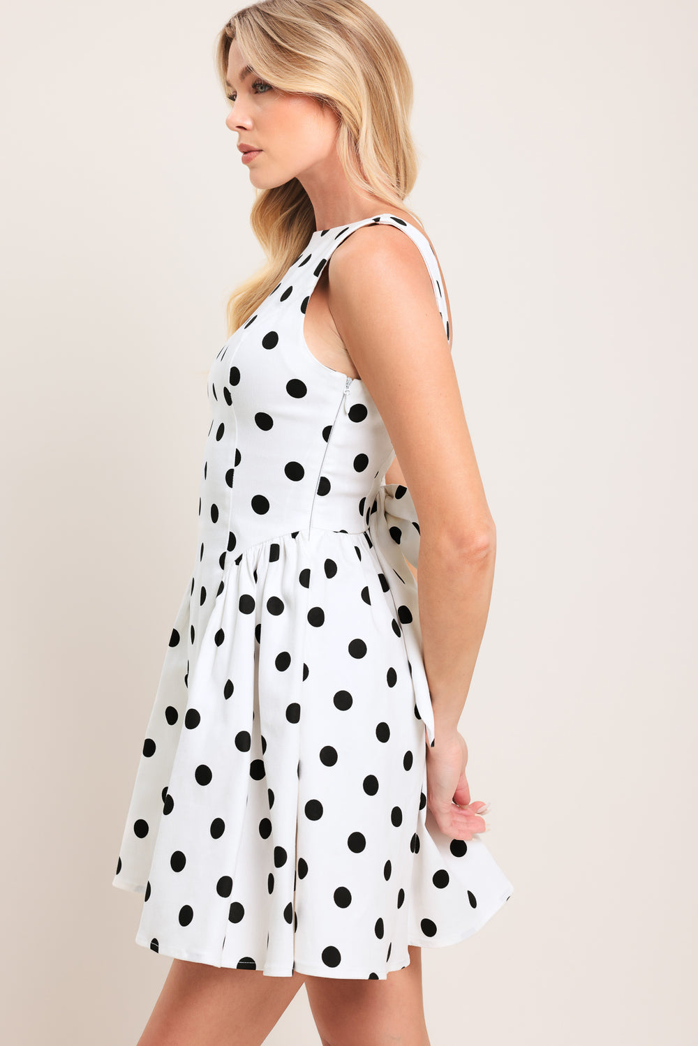 Woman wearing a sleeveless, white mini dress with black polka dots mini dress featuring boat neckline, shirred side panel, and open back with bow detail | Flying Tomato