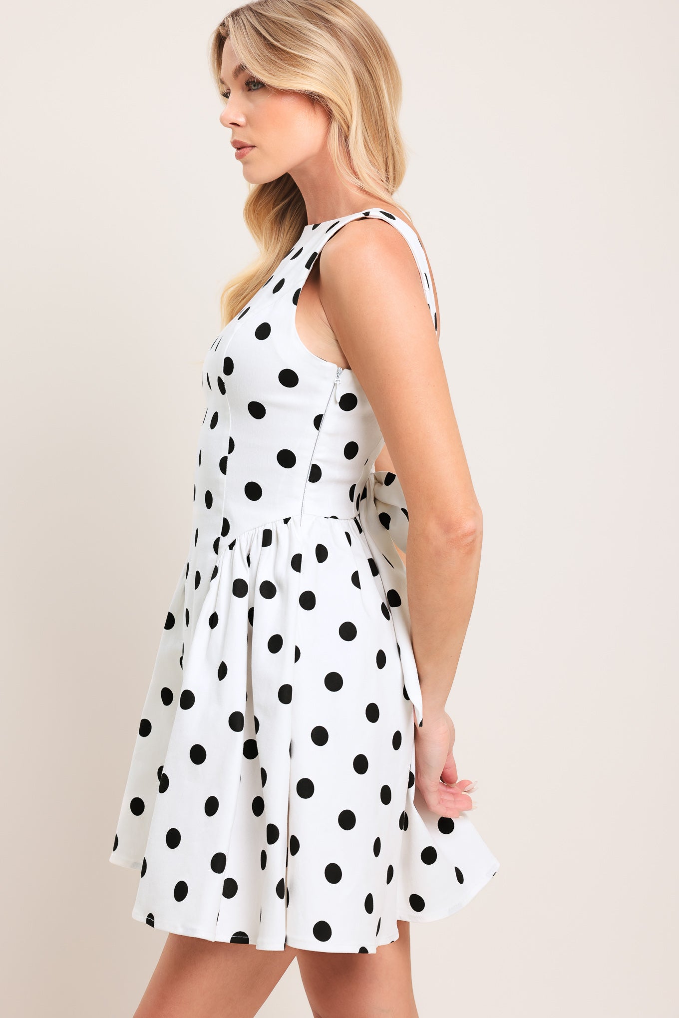 Woman wearing a sleeveless, white mini dress with black polka dots mini dress featuring boat neckline, shirred side panel, and open back with bow detail | Flying Tomato