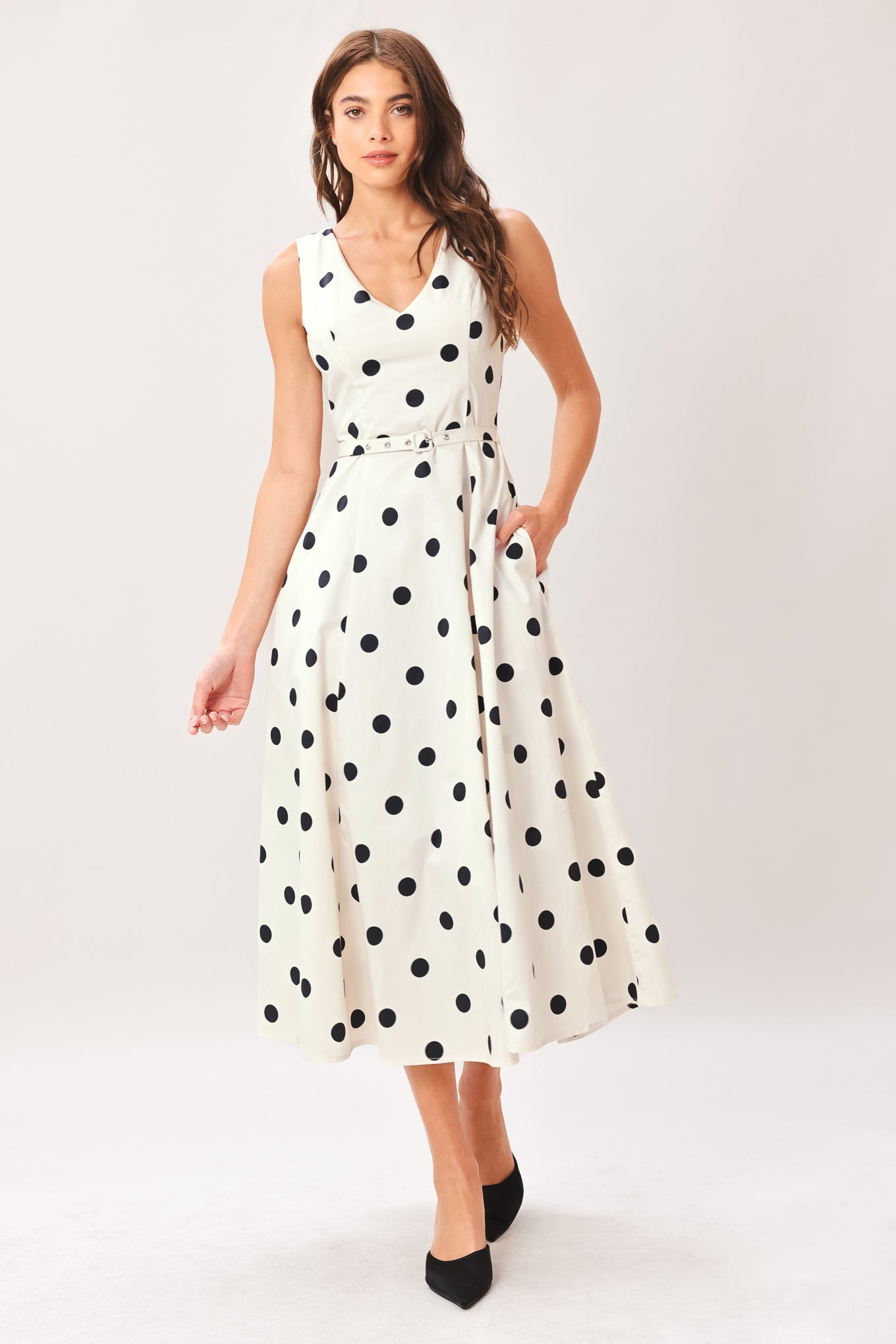 Woman wearing a white midi dress with black polka dots featuring V neckline, self belt, full circle skirt and back zipper closure | Flying Tomato