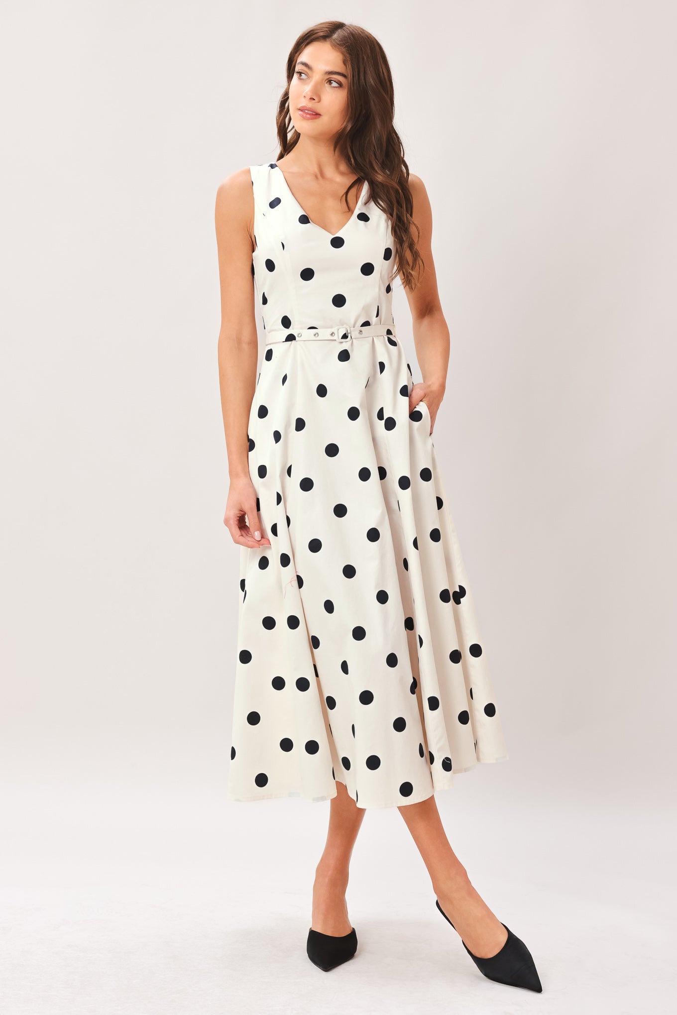 Woman wearing a white midi dress with black polka dots featuring V neckline, self belt, full circle skirt and back zipper closure | Flying Tomato