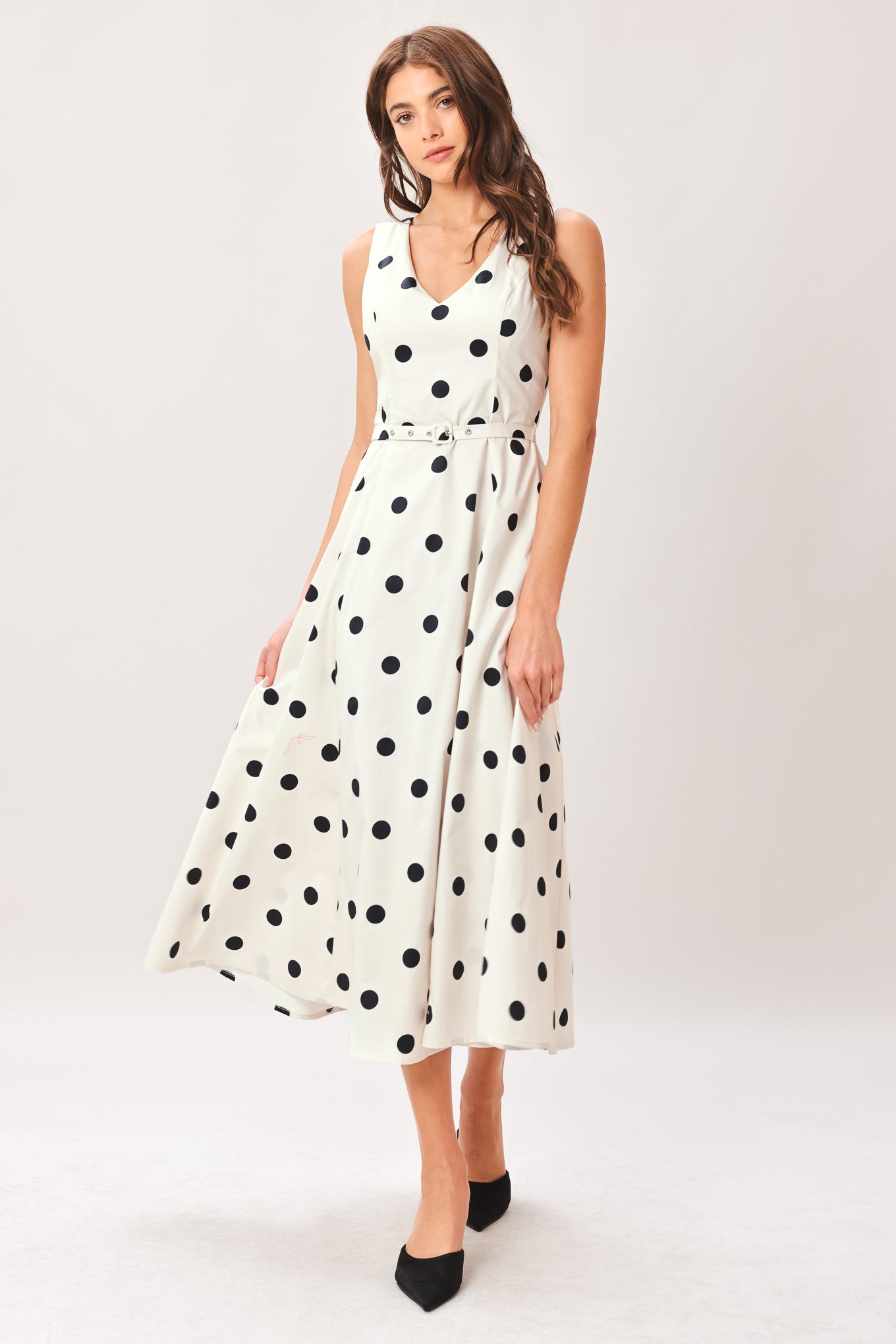 Woman wearing a white midi dress with black polka dots featuring V neckline, self belt, full circle skirt and back zipper closure | Flying Tomato