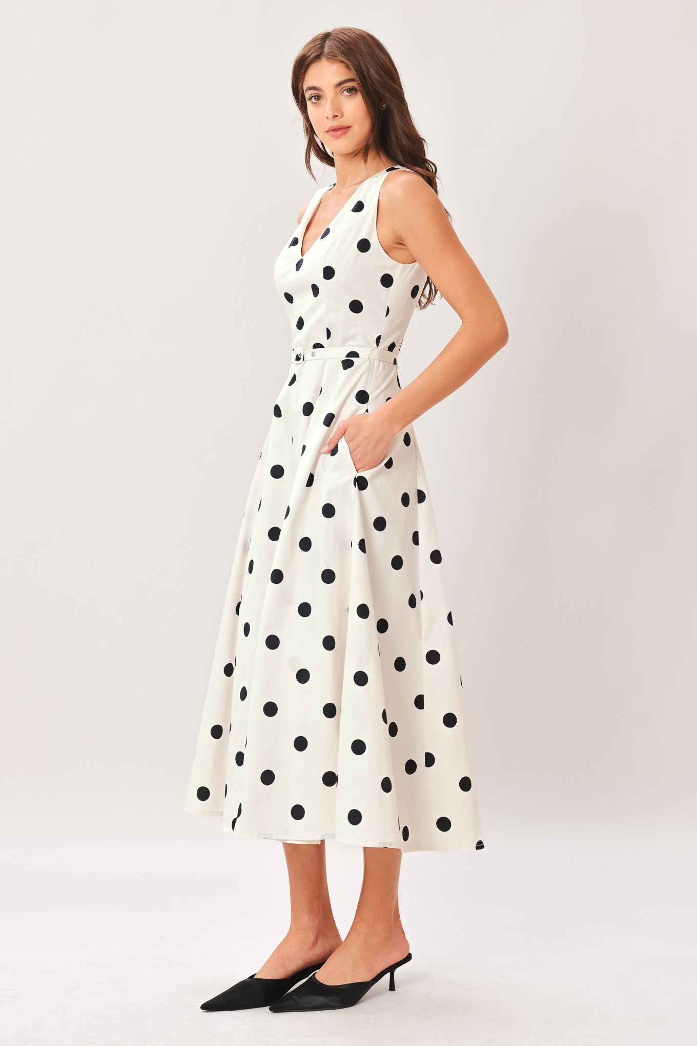 Woman wearing a white midi dress with black polka dots featuring V neckline, self belt, full circle skirt and back zipper closure | Flying Tomato