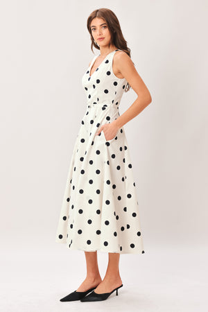 Woman wearing a white midi dress with black polka dots featuring V neckline, self belt, full circle skirt and back zipper closure | Flying Tomato