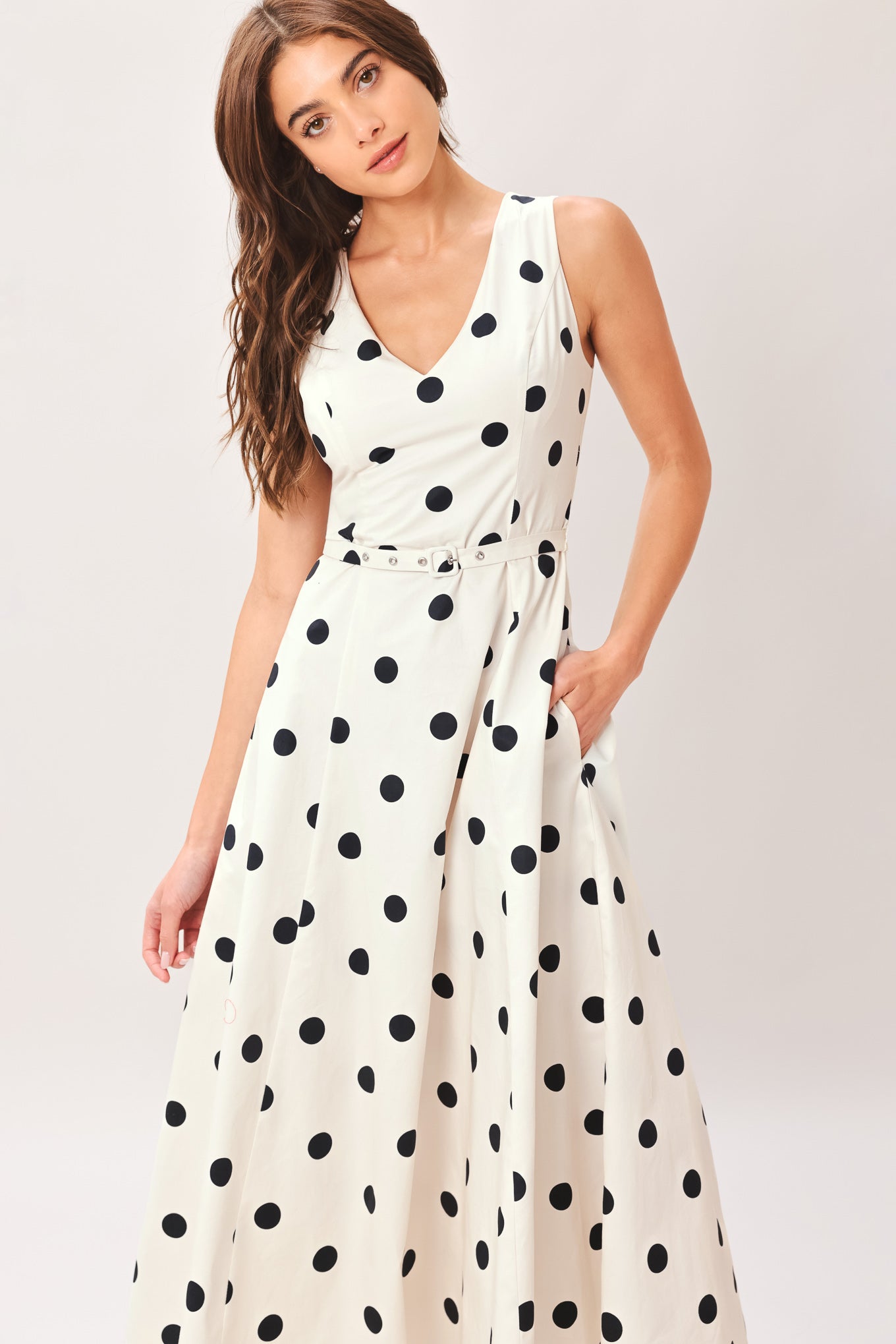 Woman wearing a white midi dress with black polka dots featuring V neckline, self belt, full circle skirt and back zipper closure | Flying Tomato