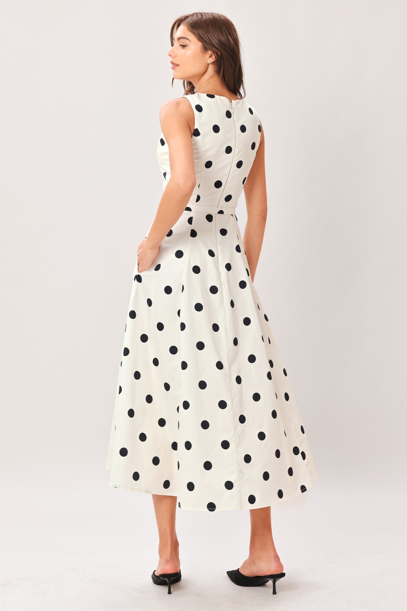 Woman wearing a white midi dress with black polka dots featuring V neckline, self belt, full circle skirt and back zipper closure | Flying Tomato