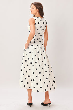 Woman wearing a white midi dress with black polka dots featuring V neckline, self belt, full circle skirt and back zipper closure | Flying Tomato