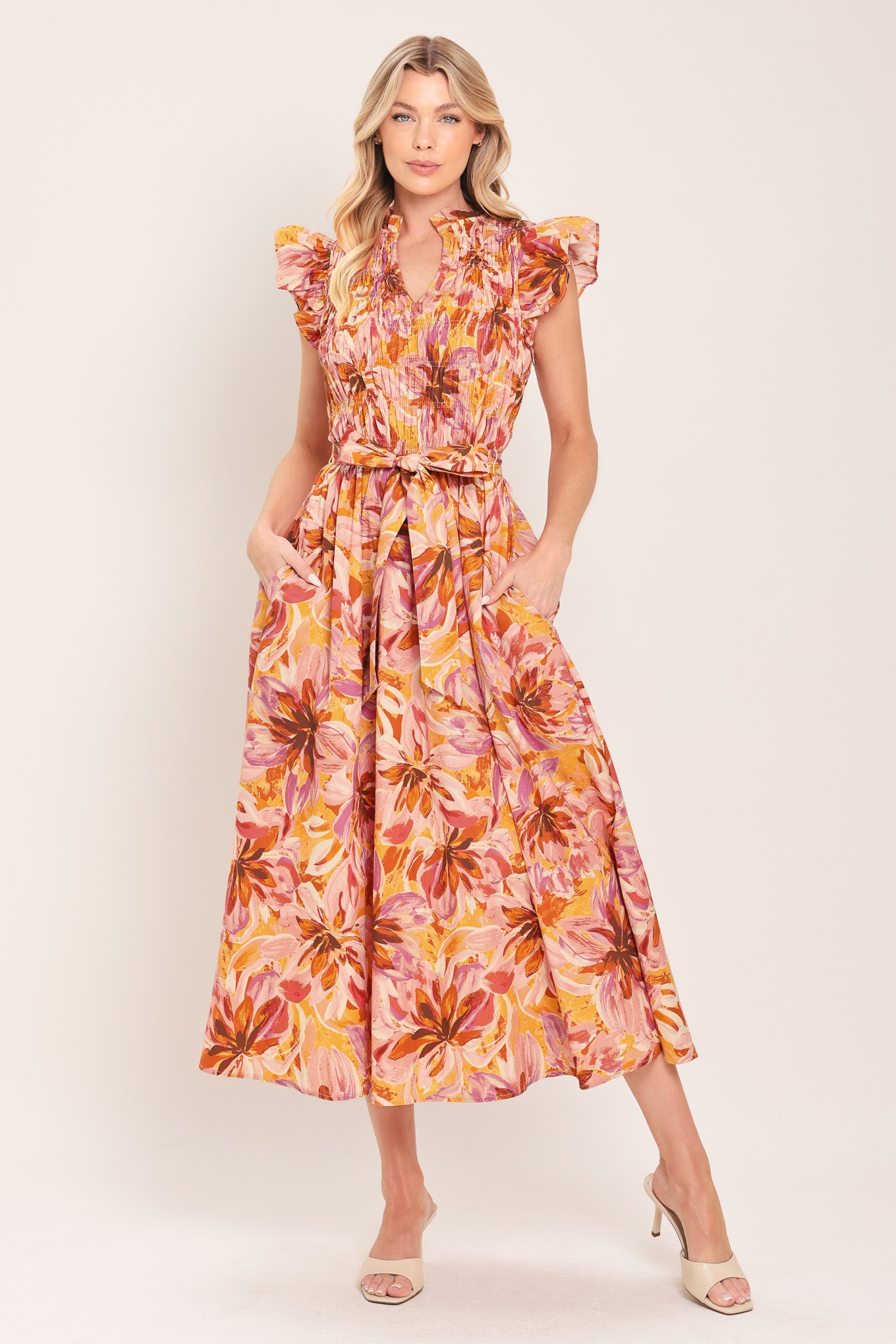 Woman wearing a pink, yellow, and orange floral printed midi dress featuring ruffle neck with V center, short ruffle sleeve, wide smocked bodice, self sash tie and full skirt | Flying Tomato