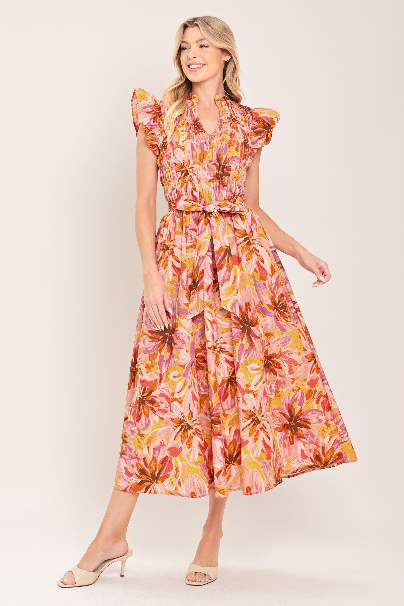 Woman wearing a pink, yellow, and orange floral printed midi dress featuring ruffle neck with V center, short ruffle sleeve, wide smocked bodice, self sash tie and full skirt | Flying Tomato