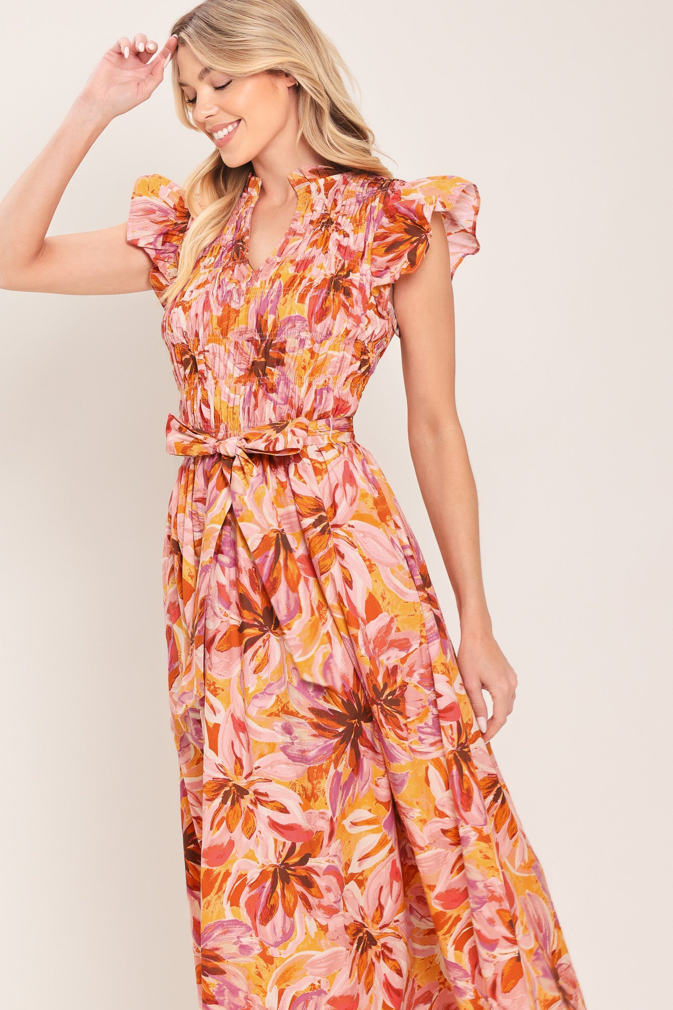 Woman wearing a pink, yellow, and orange floral printed midi dress featuring ruffle neck with V center, short ruffle sleeve, wide smocked bodice, self sash tie and full skirt | Flying Tomato