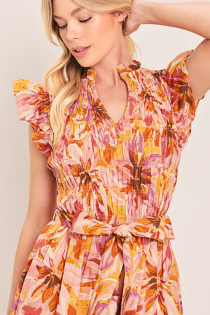 Woman wearing a pink, yellow, and orange floral printed midi dress featuring ruffle neck with V center, short ruffle sleeve, wide smocked bodice, self sash tie and full skirt | Flying Tomato