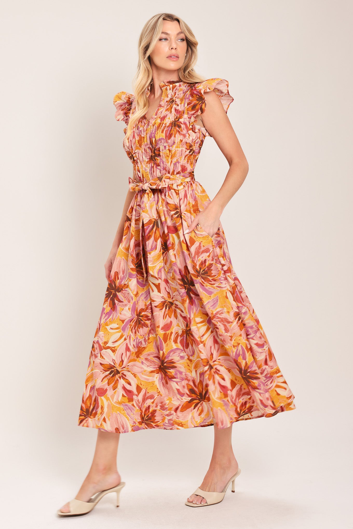 Woman wearing a pink, yellow, and orange floral printed midi dress featuring ruffle neck with V center, short ruffle sleeve, wide smocked bodice, self sash tie and full skirt | Flying Tomato