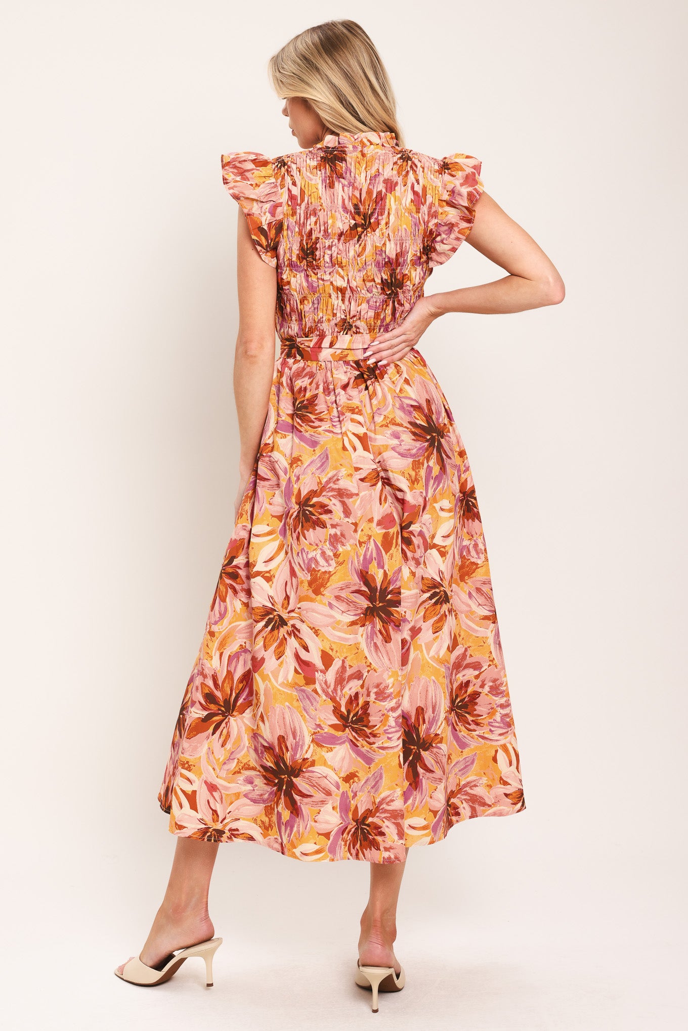 Woman wearing a pink, yellow, and orange floral printed midi dress featuring ruffle neck with V center, short ruffle sleeve, wide smocked bodice, self sash tie and full skirt | Flying Tomato