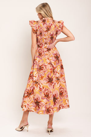 Woman wearing a pink, yellow, and orange floral printed midi dress featuring ruffle neck with V center, short ruffle sleeve, wide smocked bodice, self sash tie and full skirt | Flying Tomato