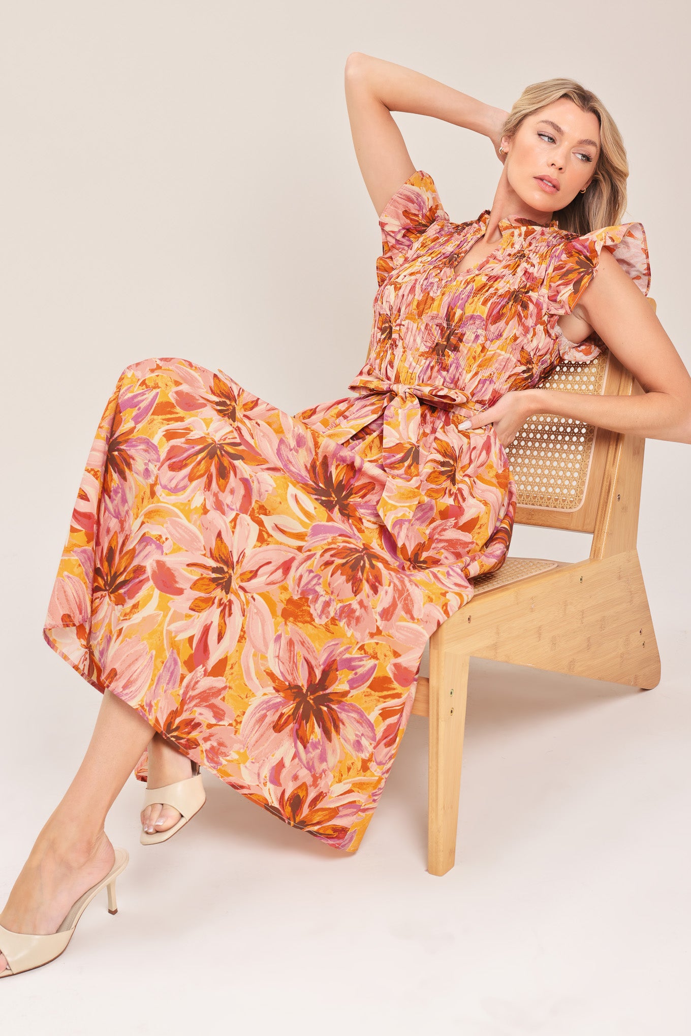 Woman wearing a pink, yellow, and orange floral printed midi dress featuring ruffle neck with V center, short ruffle sleeve, wide smocked bodice, self sash tie and full skirt | Flying Tomato