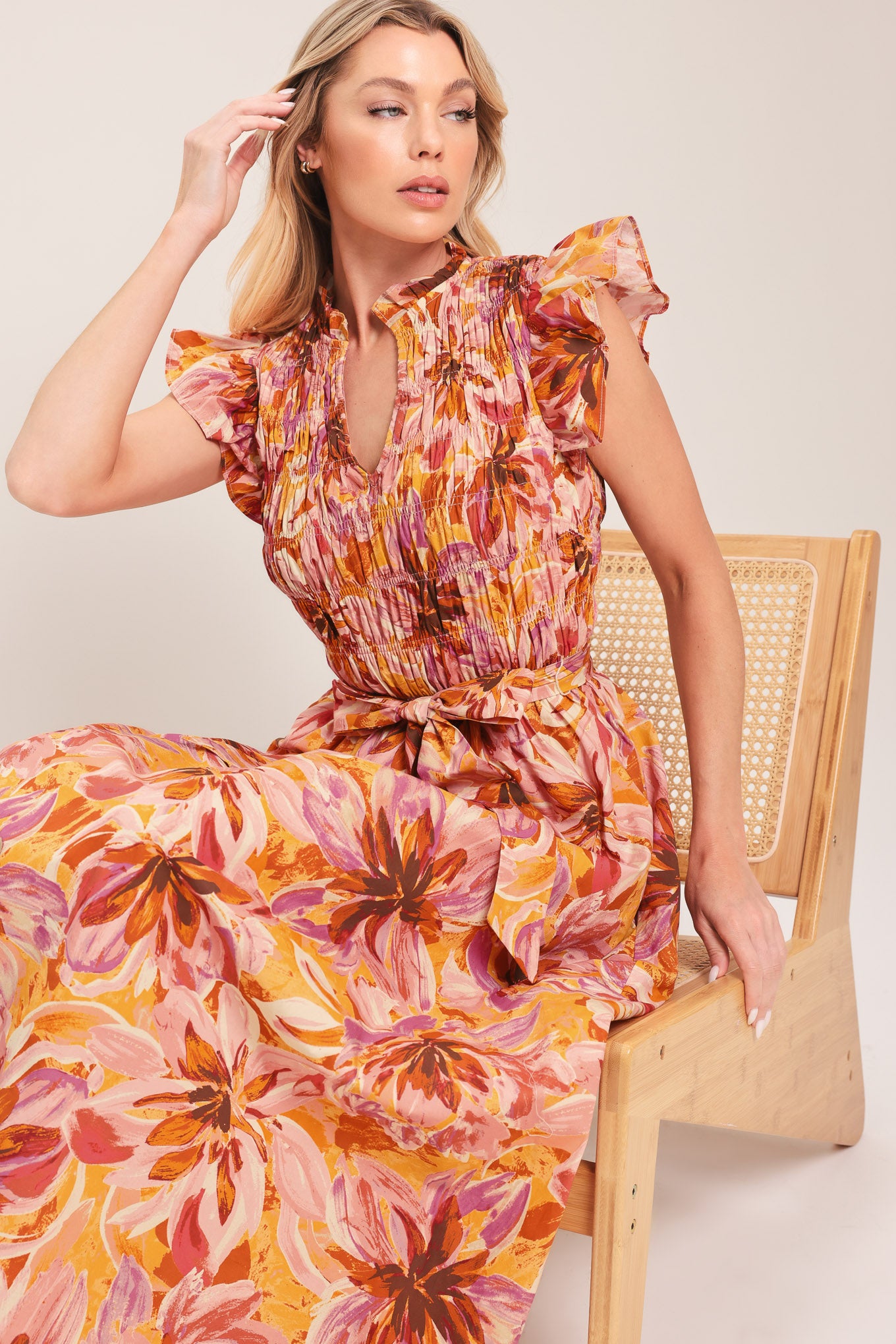 Woman wearing a pink, yellow, and orange floral printed midi dress featuring ruffle neck with V center, short ruffle sleeve, wide smocked bodice, self sash tie and full skirt | Flying Tomato