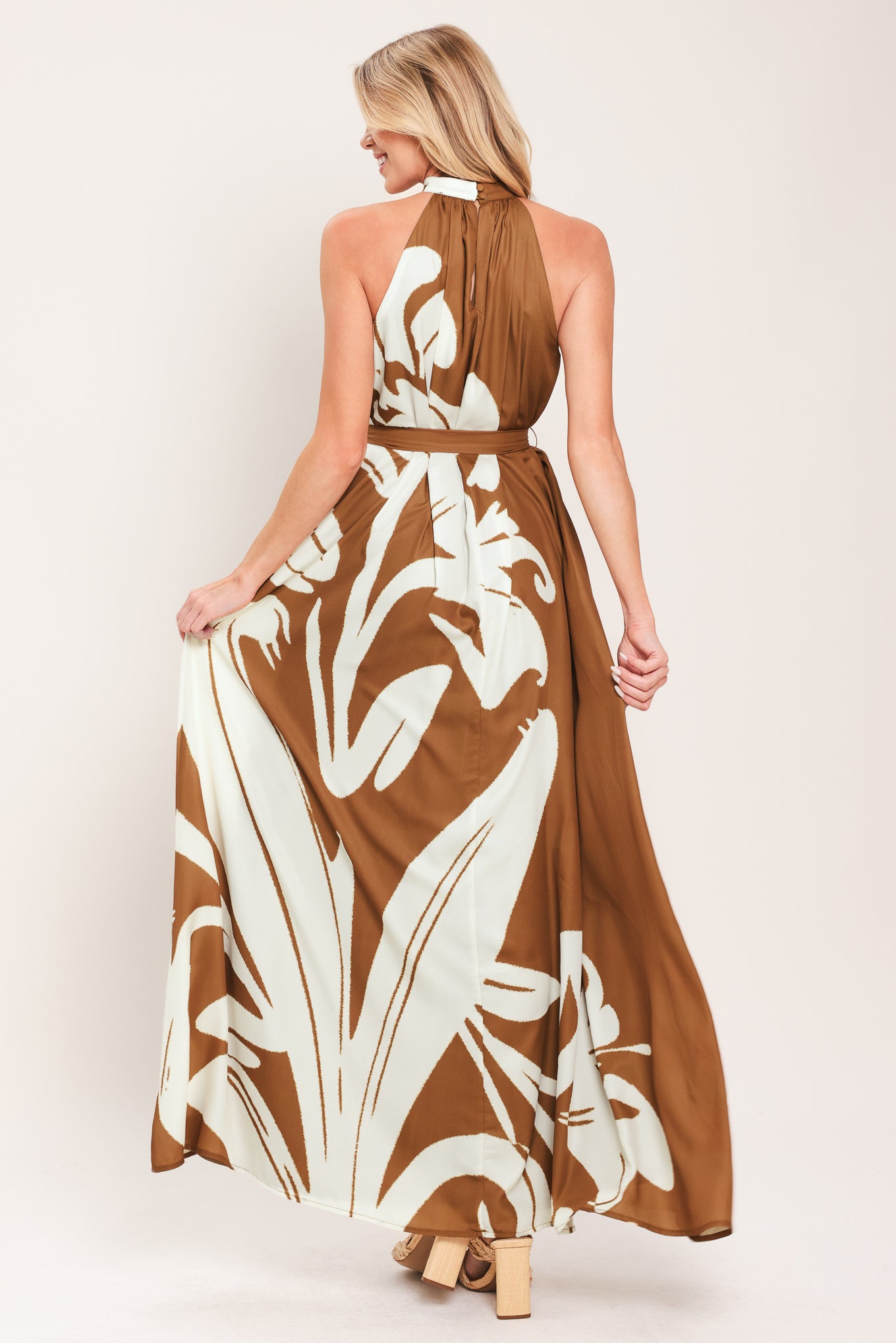Woman wearing a brown maxi dress featuring halter neckline with back neck button closure, sleeveless and self sash tie | Flying Tomato