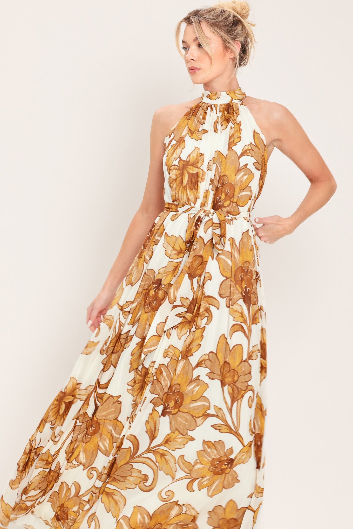 Woman wearing an ivory with brown floral print maxi dress with halter neckline with tie back, pleated body and self sash tie | Flying Tomato