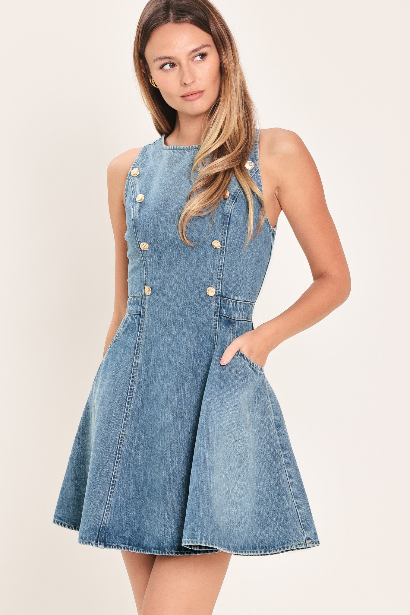 Woman wearing a mini denim dress featuring round neckline, sleeveless, front button down detail, side pockets, circle skirt and back zipper closure | Flying Tomato