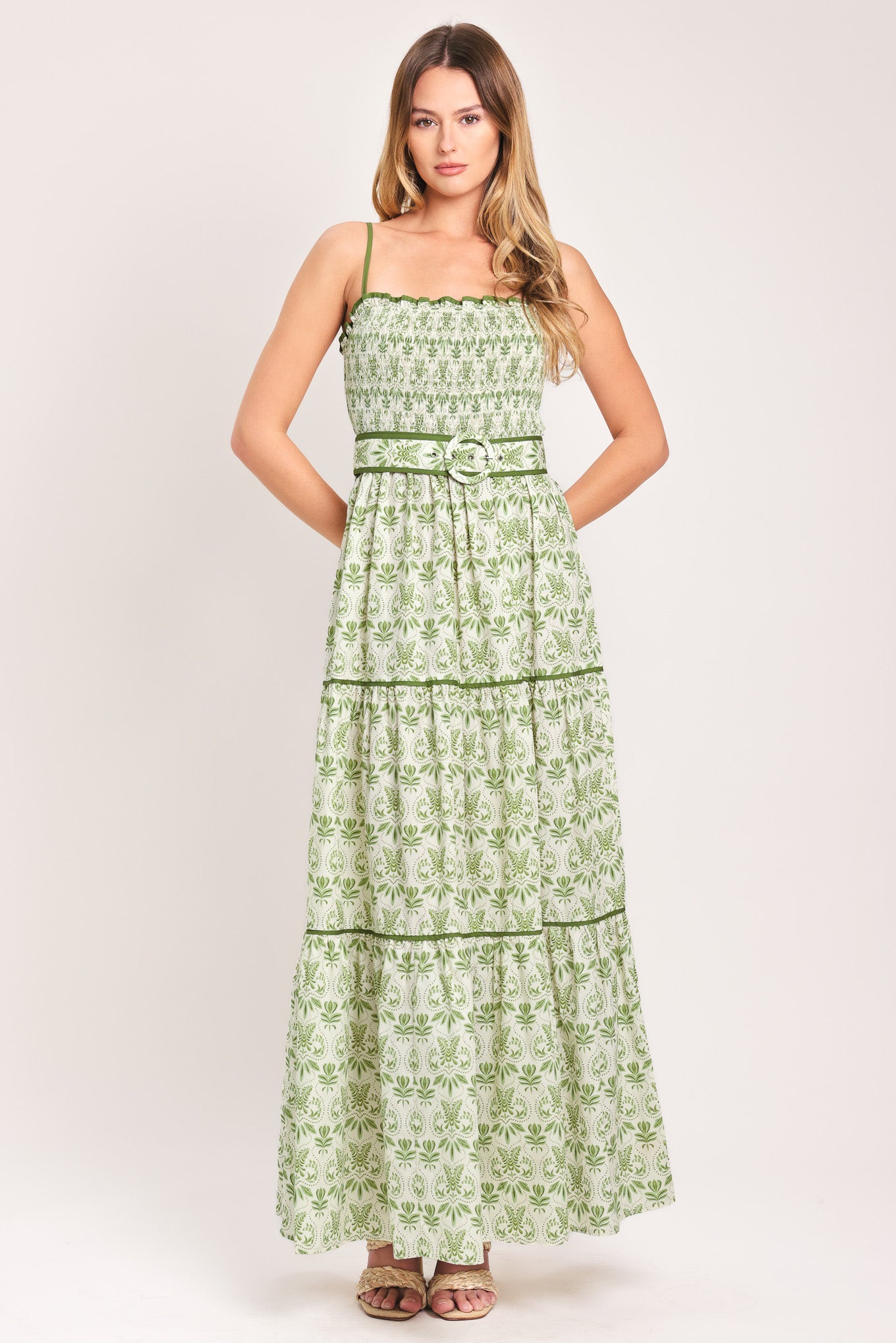 Woman wearing a green floral maxi dress featuring straight neckline with ruffle edge, straps, smocked bodice, self belt and tier skirt with piping inset | Flying Tomato