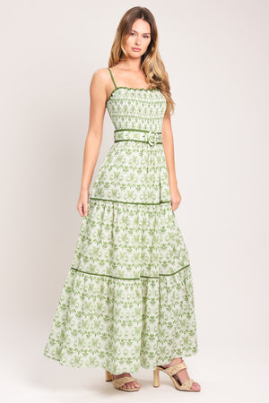 Woman wearing a green floral maxi dress featuring straight neckline with ruffle edge, straps, smocked bodice, self belt and tier skirt with piping inset | Flying Tomato