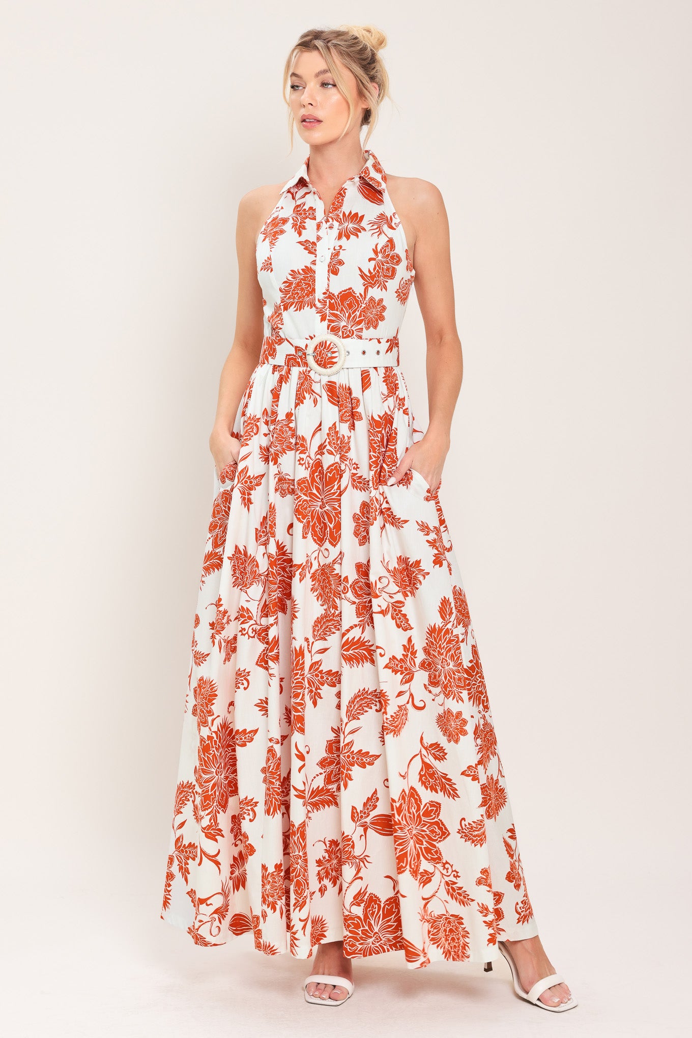 Woman wearing an ivory with rust-colored floral pattern maxi dress featuring halter neckline with collar, self belt, side pockets and full skirt | Flying Tomato
