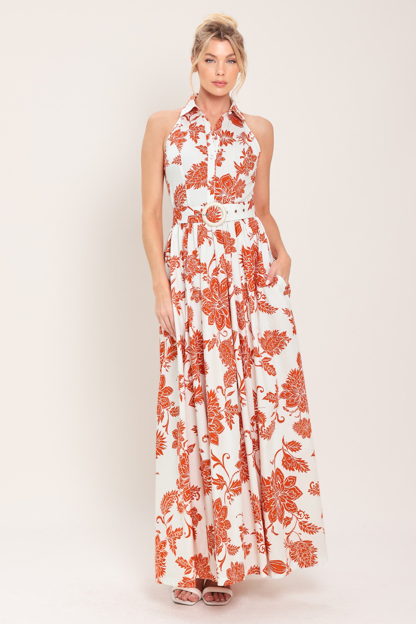 Woman wearing an ivory with rust-colored floral pattern maxi dress featuring halter neckline with collar, self belt, side pockets and full skirt | Flying Tomato