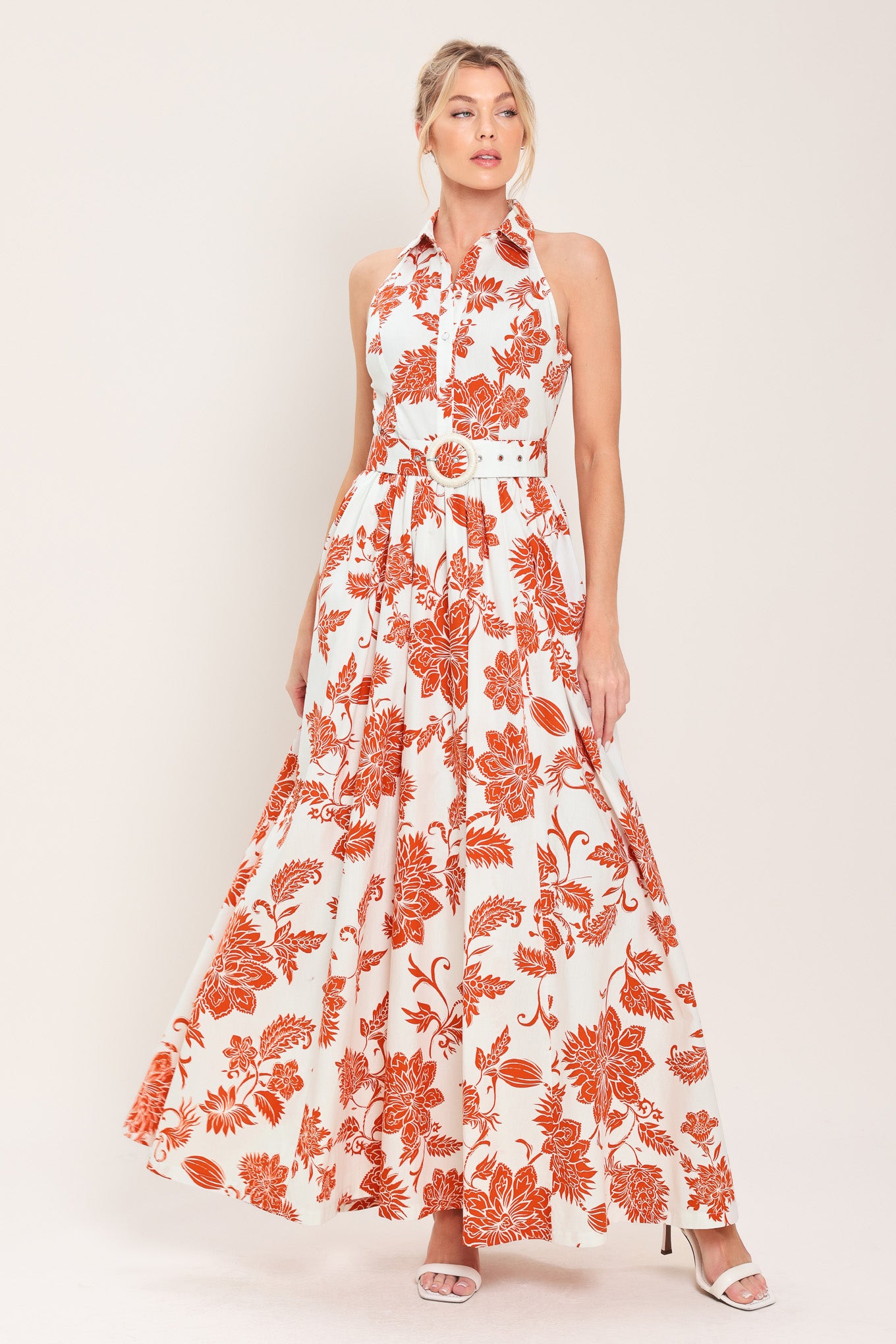 Woman wearing an ivory with rust-colored flowers and leaves maxi dress featuring halter neckline with collar, self belt, side pockets and full skirt | Flying Tomato