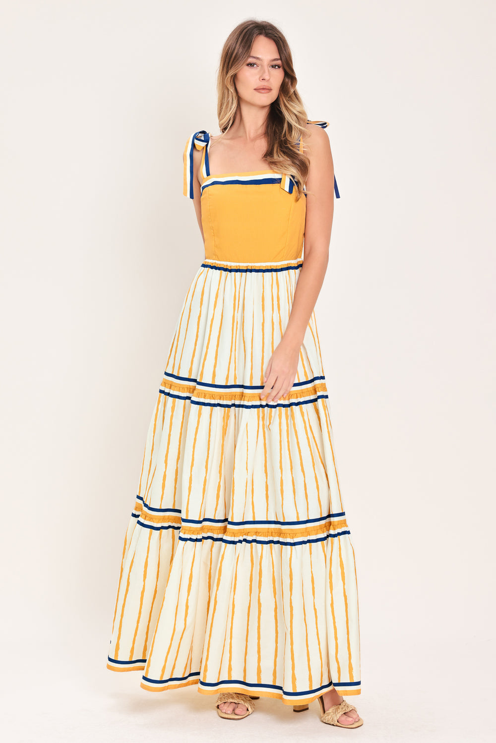 Woman wearing a camel yellow and blue striped maxi dress featuring straight neckline, shoulder ties, tier skirt and smocked back bodice | Flying Tomato