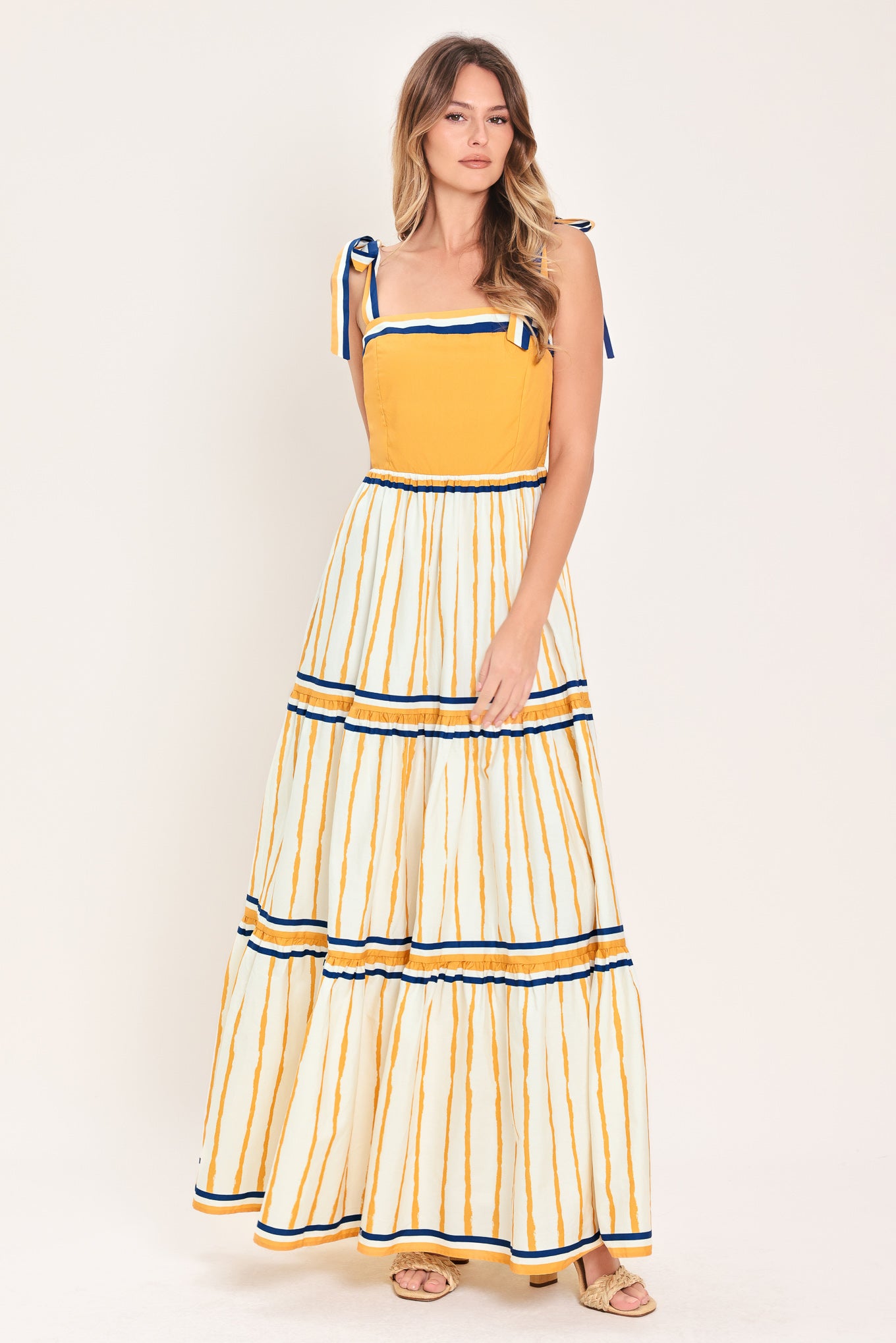 Woman wearing a camel yellow and blue striped maxi dress featuring straight neckline, shoulder ties, tier skirt and smocked back bodice | Flying Tomato