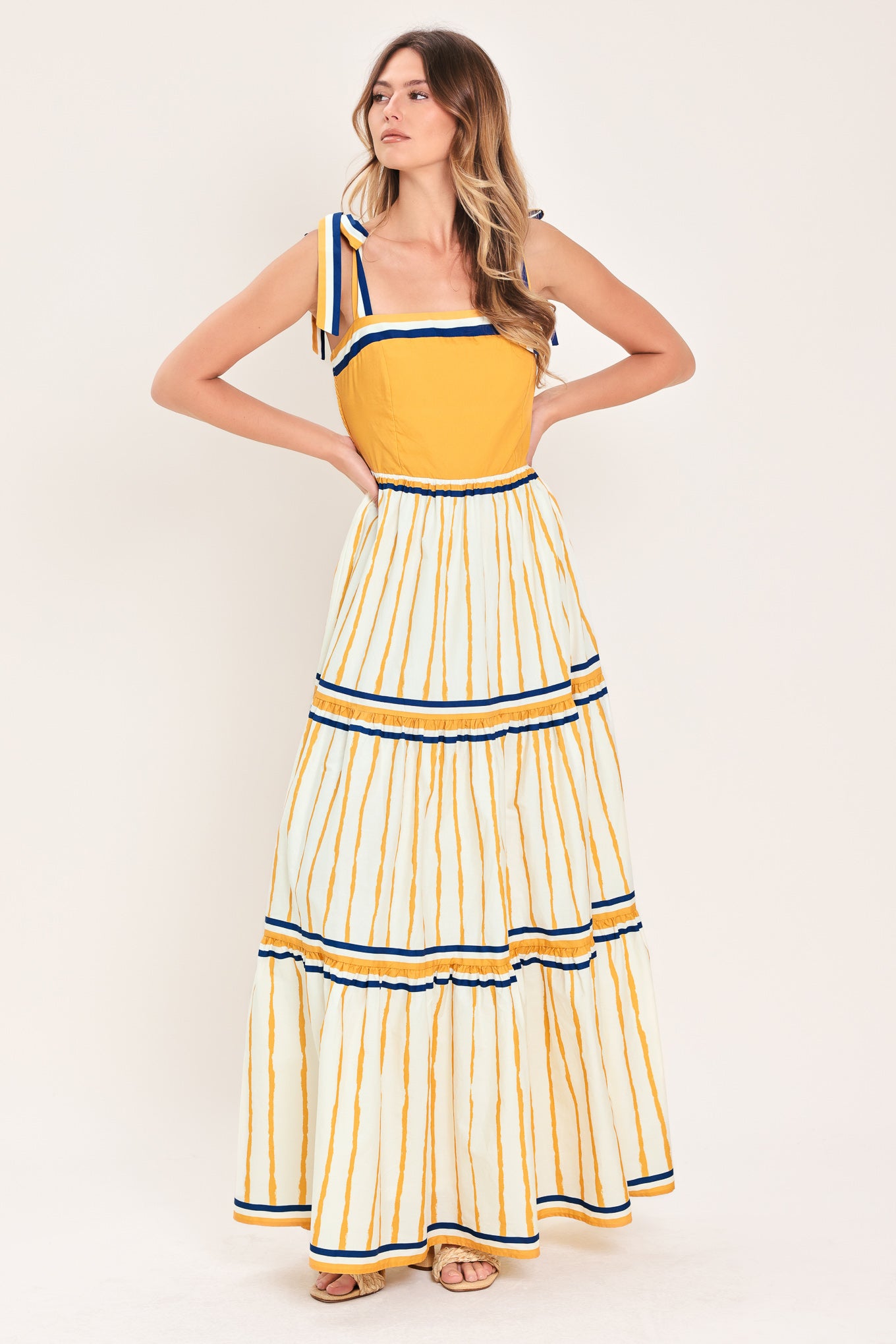 Woman wearing a camel yellow and blue striped maxi dress featuring straight neckline, shoulder ties, tier skirt and smocked back bodice | Flying Tomato