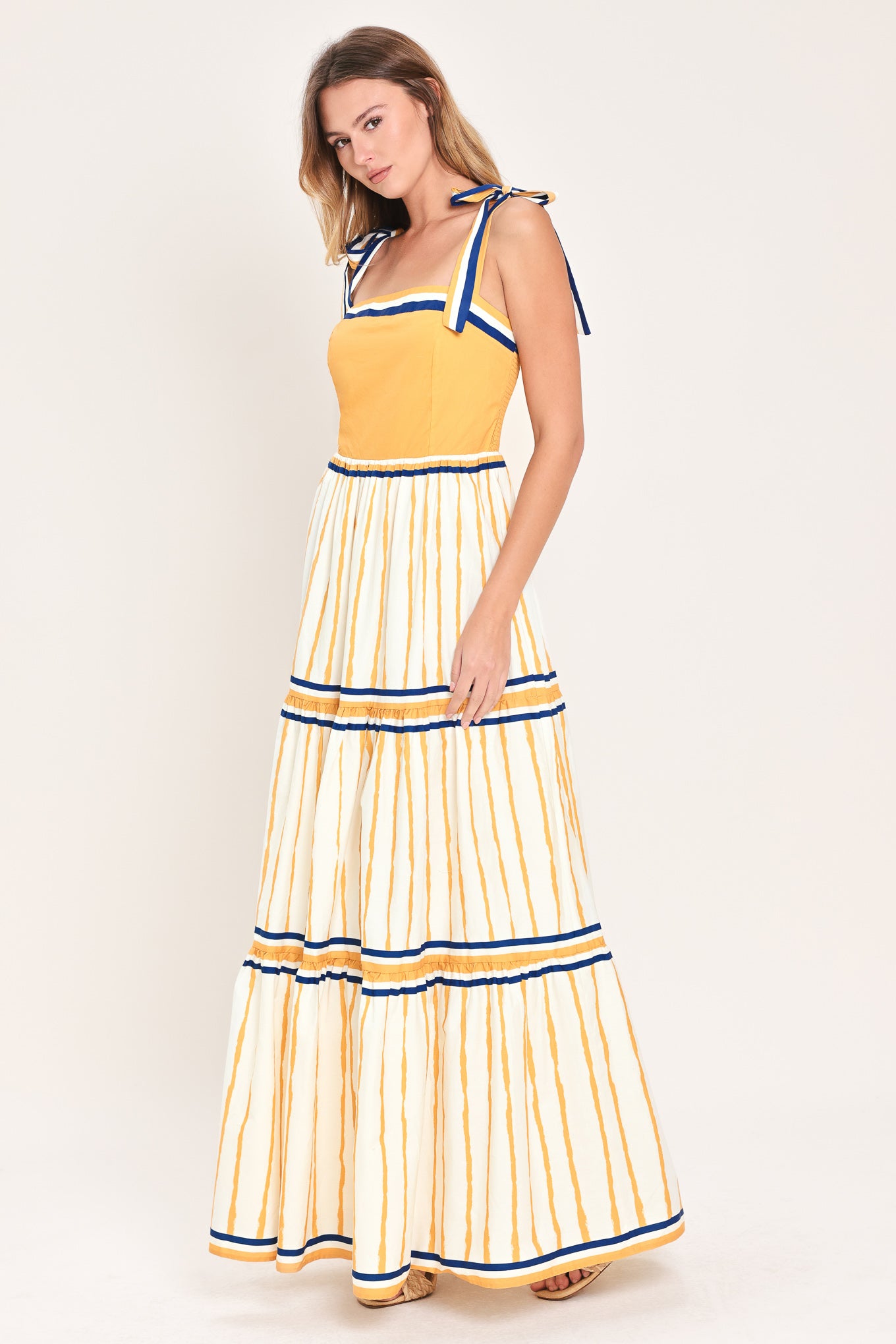 Woman wearing a camel yellow and blue striped maxi dress featuring straight neckline, shoulder ties, tier skirt and smocked back bodice | Flying Tomato