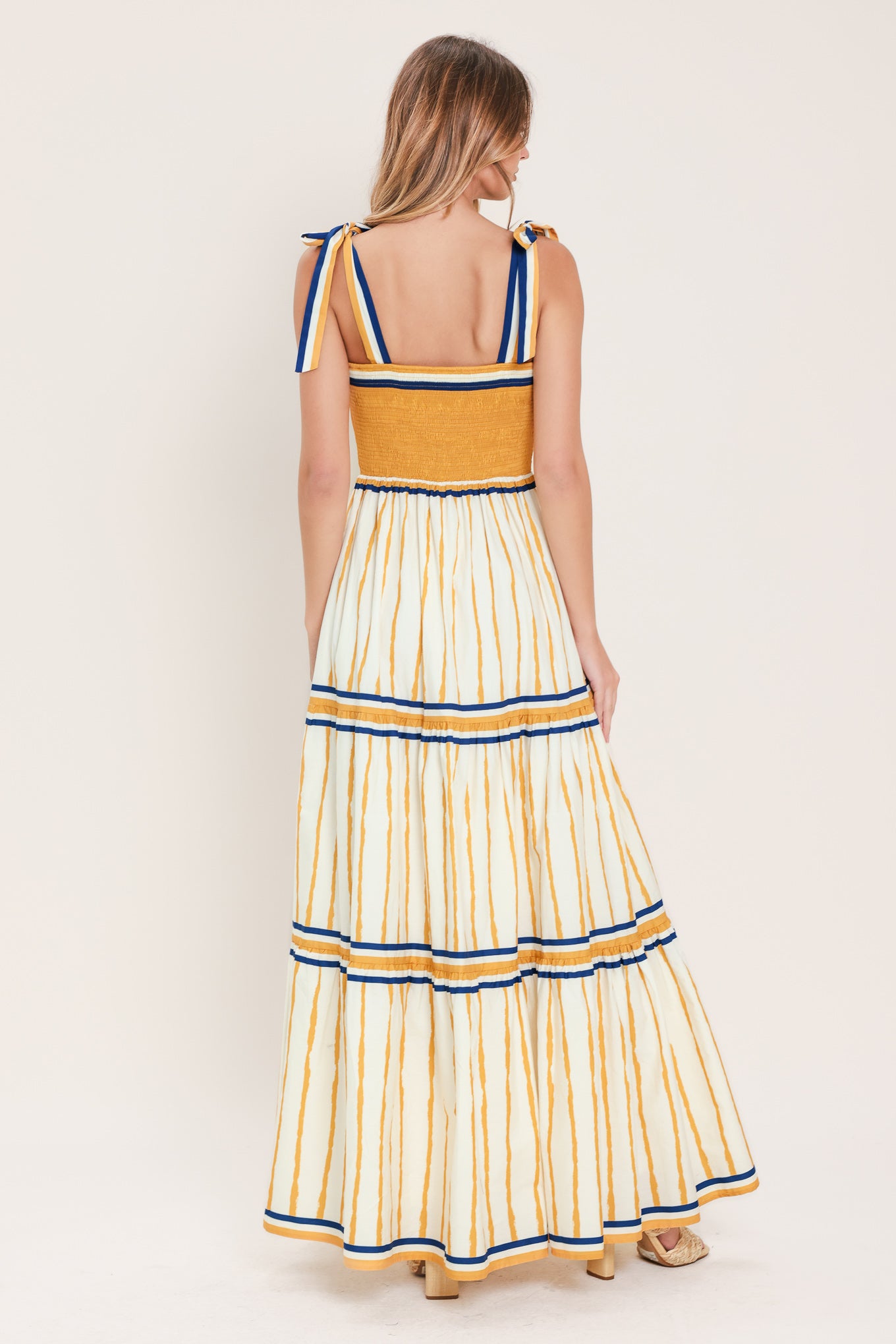 Woman wearing a camel yellow and blue striped maxi dress featuring straight neckline, shoulder ties, tier skirt and smocked back bodice | Flying Tomato
