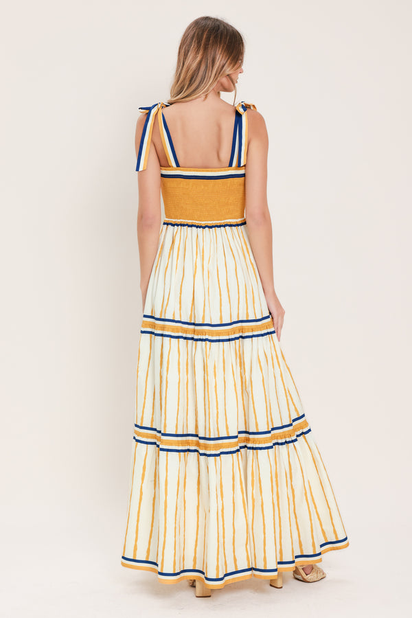 Woman wearing a camel yellow and blue striped maxi dress featuring straight neckline, shoulder ties, tier skirt and smocked back bodice | Flying Tomato