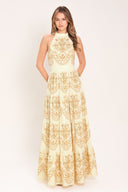 Woman wearing an off white with gold printed details maxi dress featuring halter neckline with back tie, tier skirt and back zipper closure | Flying Tomato
