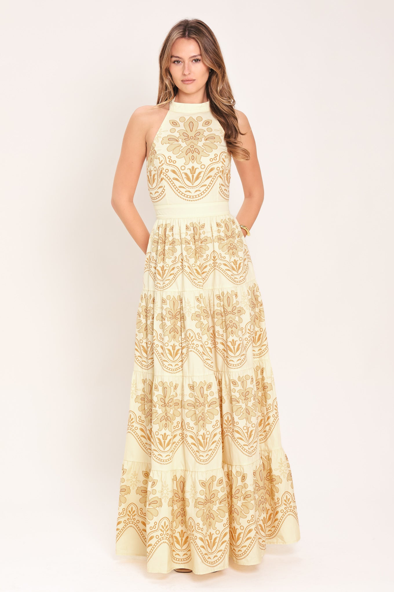 Woman wearing an off white with gold printed details maxi dress featuring halter neckline with back tie, tier skirt and back zipper closure | Flying Tomato