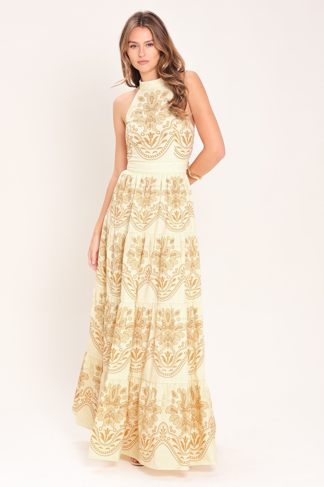 Woman wearing an off white with gold printed details maxi dress featuring halter neckline with back tie, tier skirt and back zipper closure | Flying Tomato