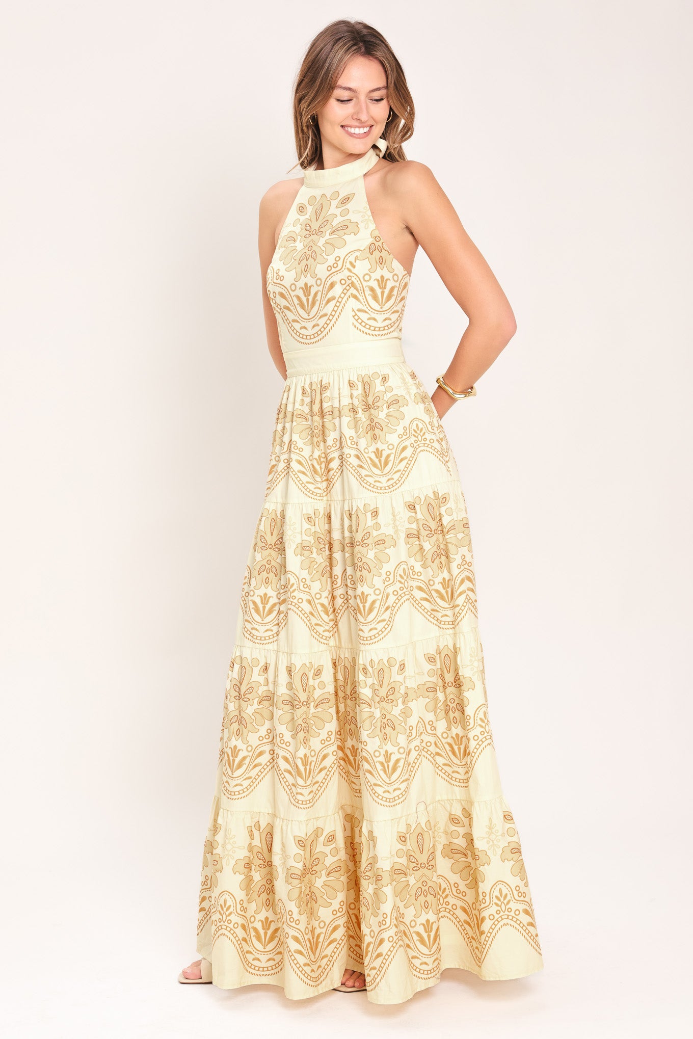 Woman wearing an off white with gold printed details maxi dress featuring halter neckline with back tie, tier skirt and back zipper closure | Flying Tomato