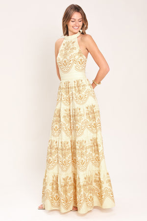 Woman wearing an off white with gold printed details maxi dress featuring halter neckline with back tie, tier skirt and back zipper closure | Flying Tomato