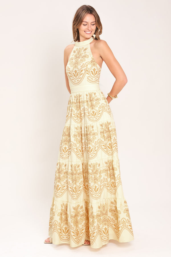 Woman wearing an off white with gold printed details maxi dress featuring halter neckline with back tie, tier skirt and back zipper closure | Flying Tomato