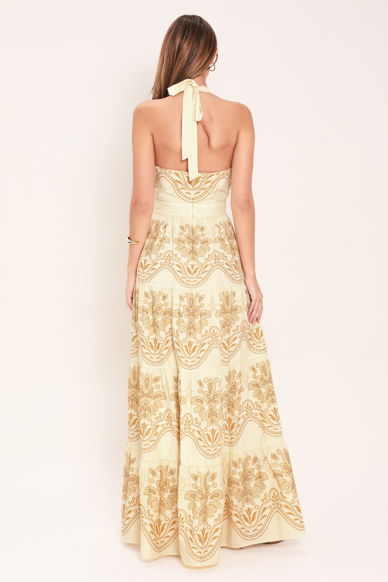 Woman wearing an off white with gold printed details maxi dress featuring halter neckline with back tie, tier skirt and back zipper closure | Flying Tomato