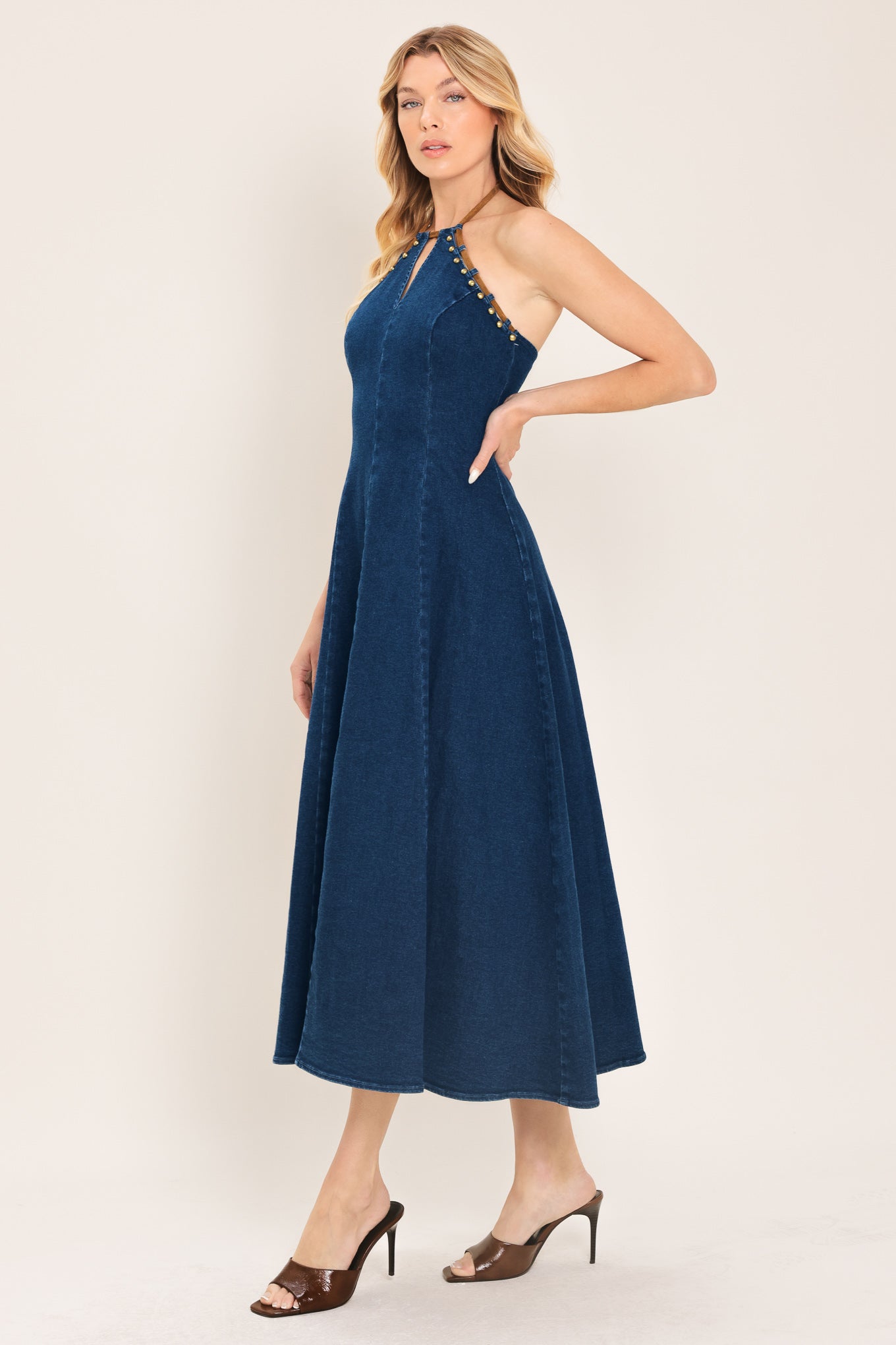 Woman wearing a blue denim midi dress featuring halter neckline with contrasting tie, stud detail, circle skirt and back zipper closure | Flying Tomato