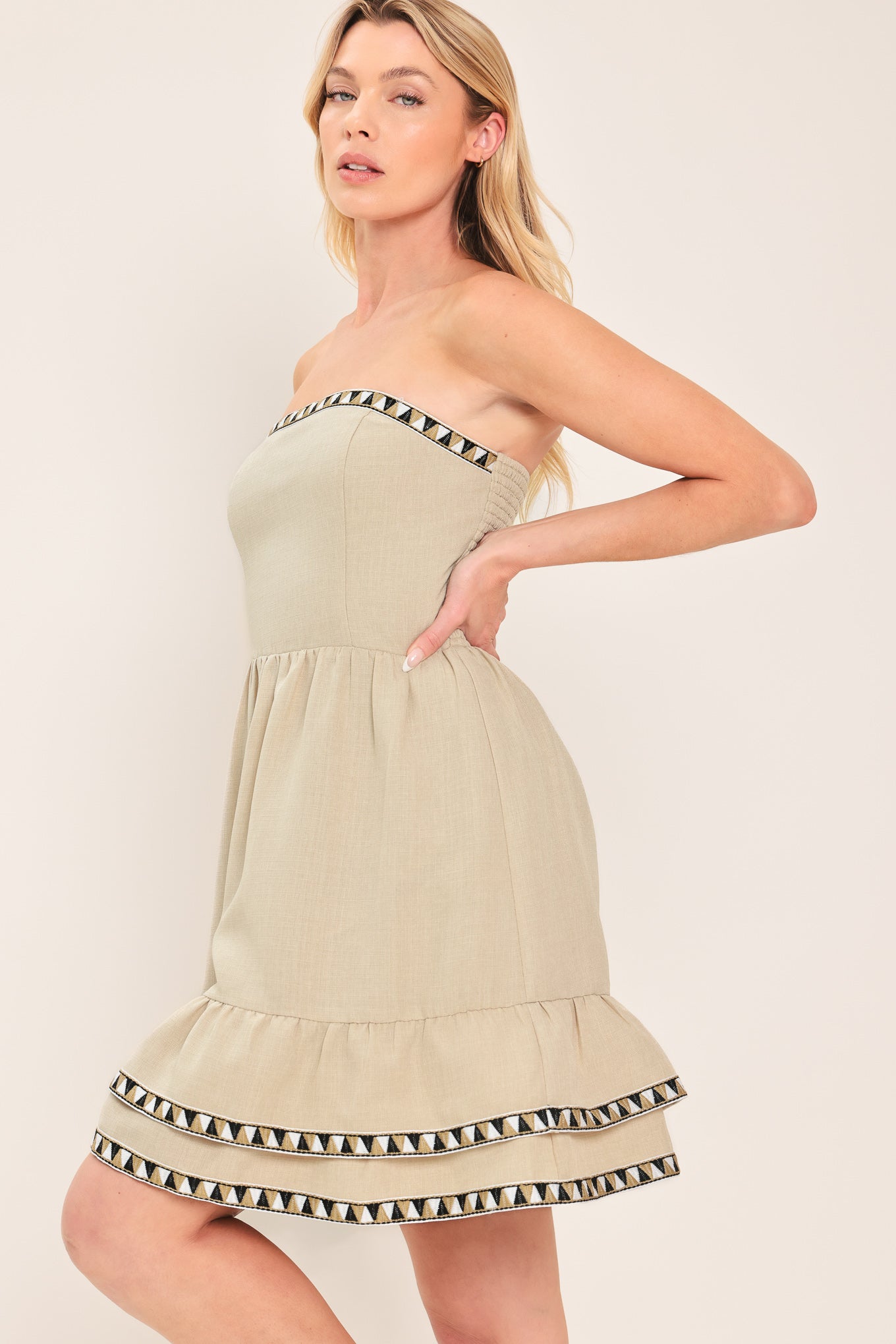 Woman wearing a stone beige strapless mini dress featuring double ruffle hem with embroidered edge and smocked back bodice | Flying Tomato