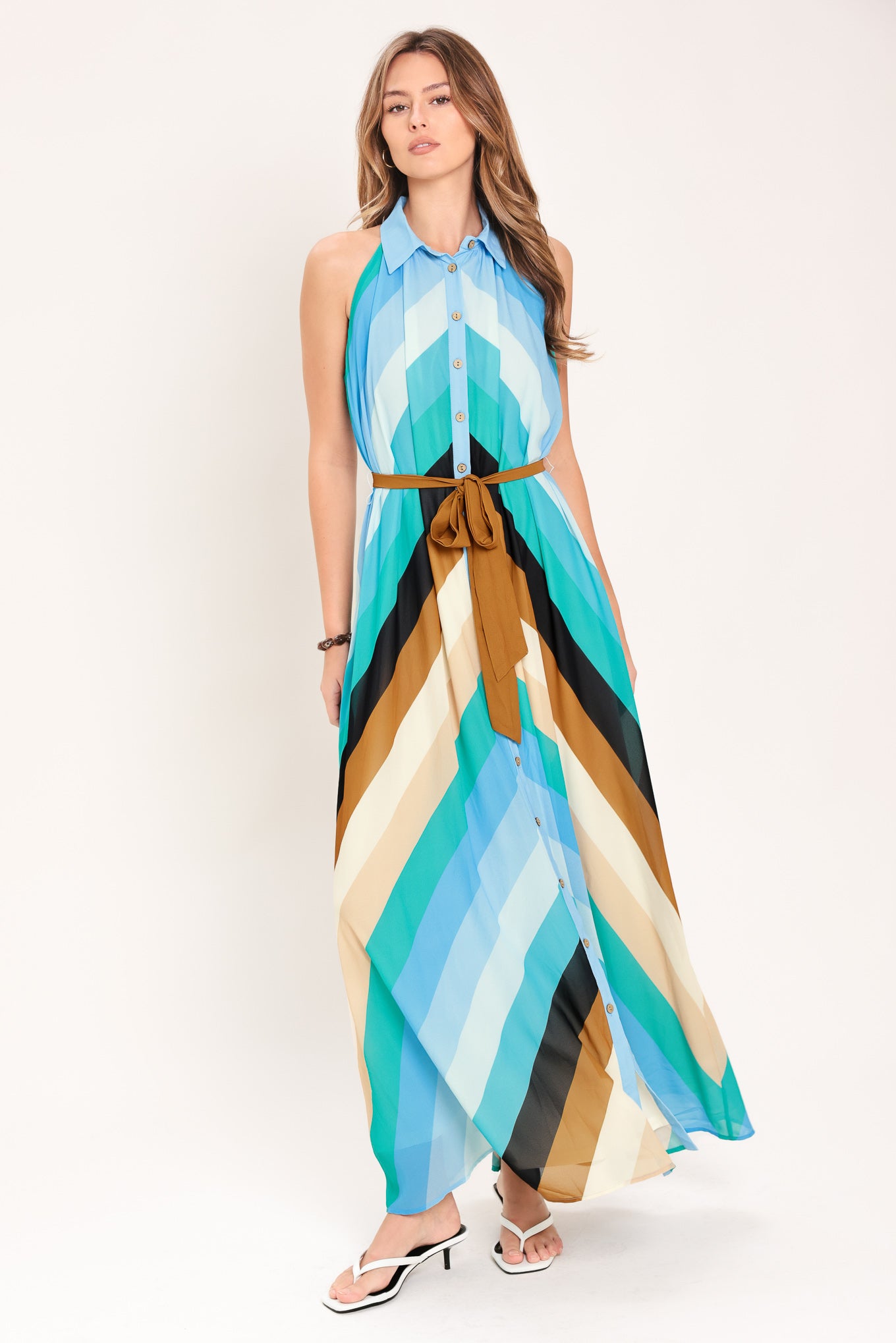 Woman wearing a sleeveless multicolor blue, teal, and brown striped maxi dress featuring halter neckline with collared finish, contrasting belt, and front button down | Flying Tomato