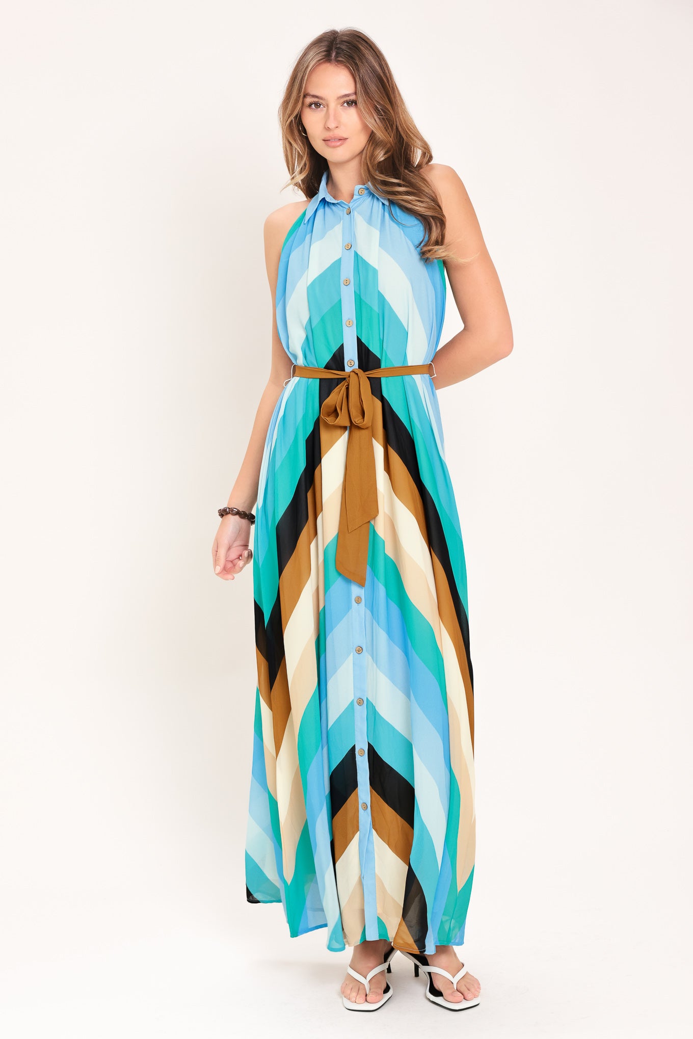 Woman wearing a sleeveless multicolor blue, teal, and brown striped maxi dress featuring halter neckline with collared finish, contrasting belt, and front button down | Flying Tomato
