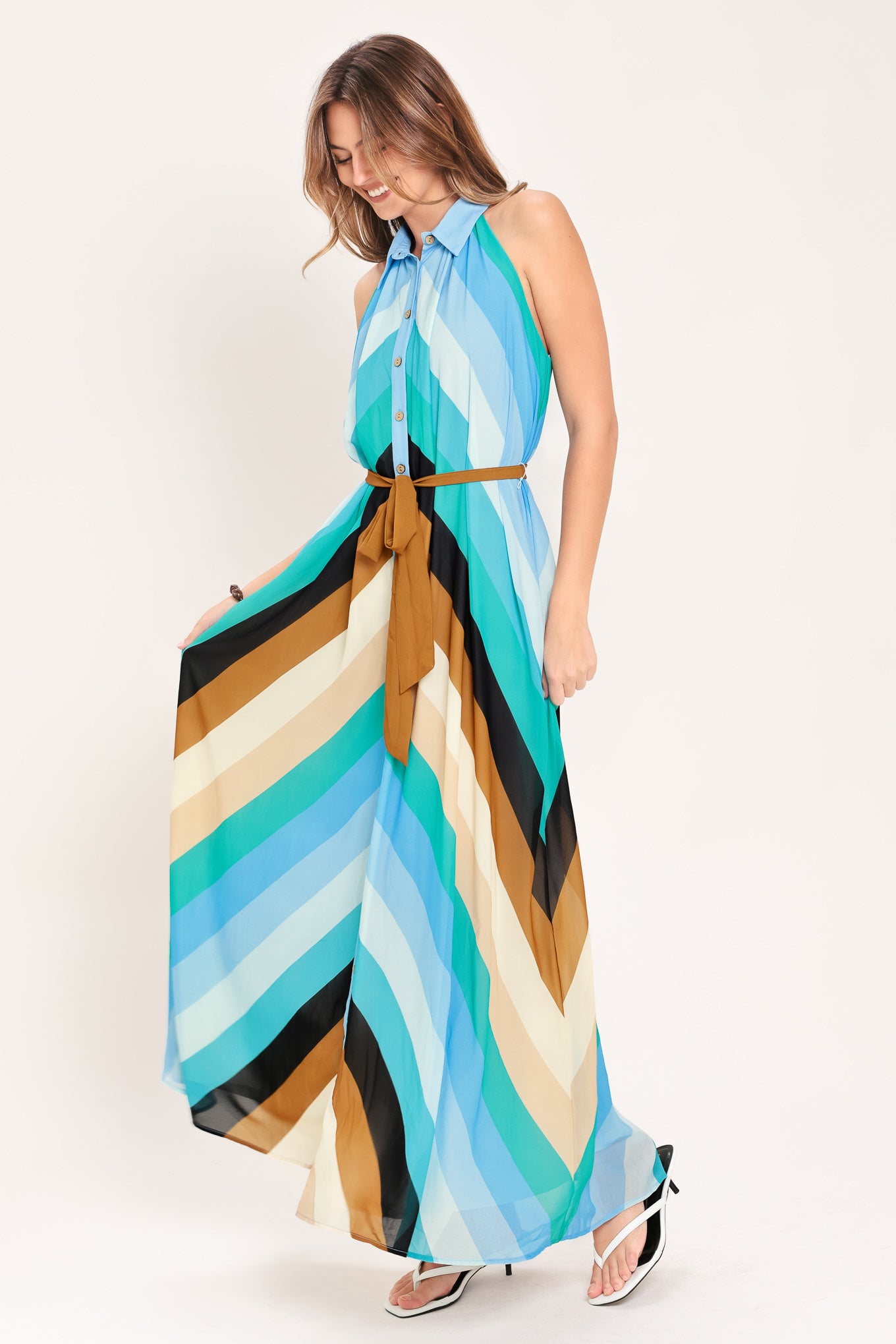 Woman wearing a sleeveless multicolor blue, teal, and brown striped maxi dress featuring halter neckline with collared finish, contrasting belt, and front button down | Flying Tomato