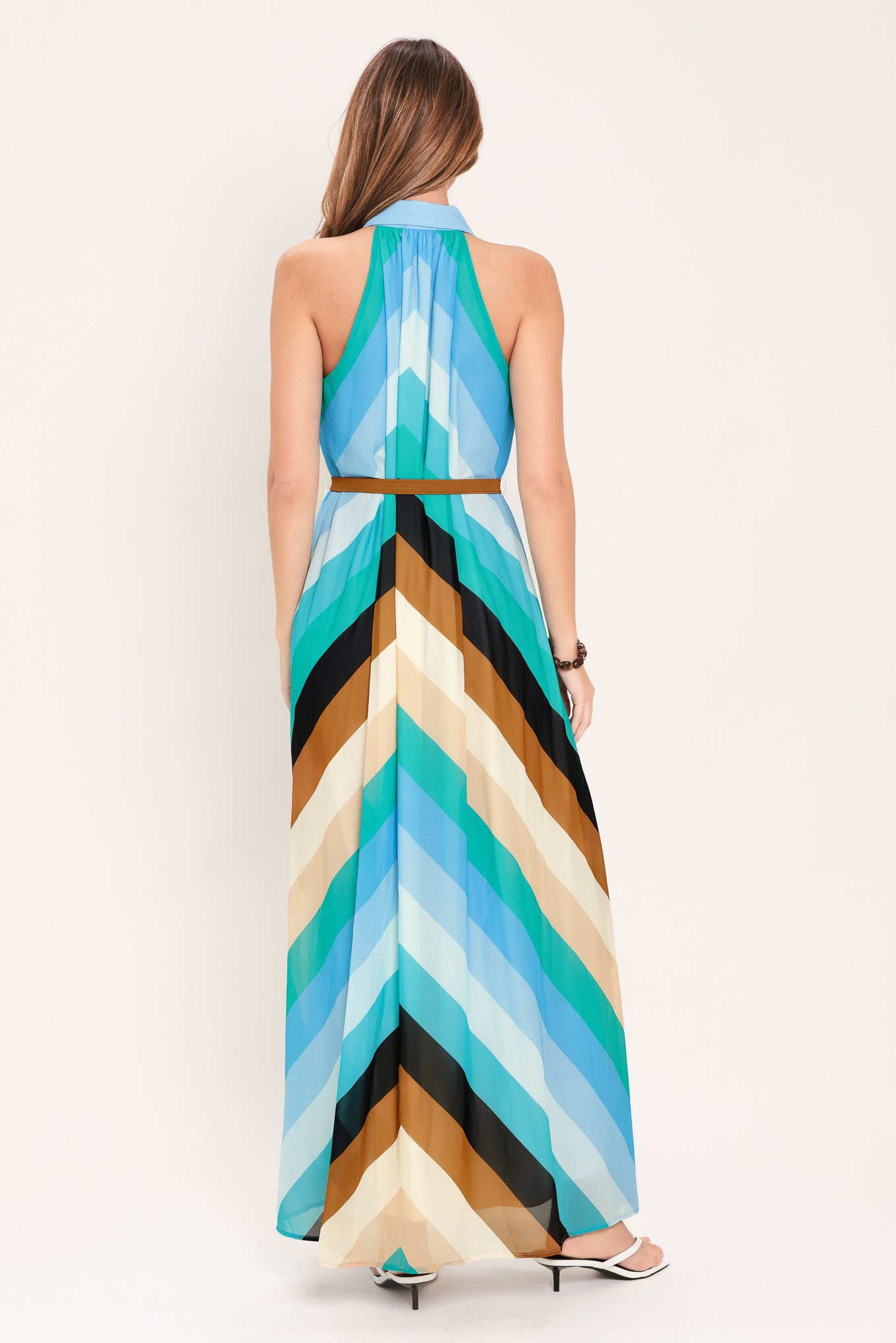 Woman wearing a sleeveless multicolor blue, teal, and brown striped maxi dress featuring halter neckline with collared finish, contrasting belt, and front button down | Flying Tomato