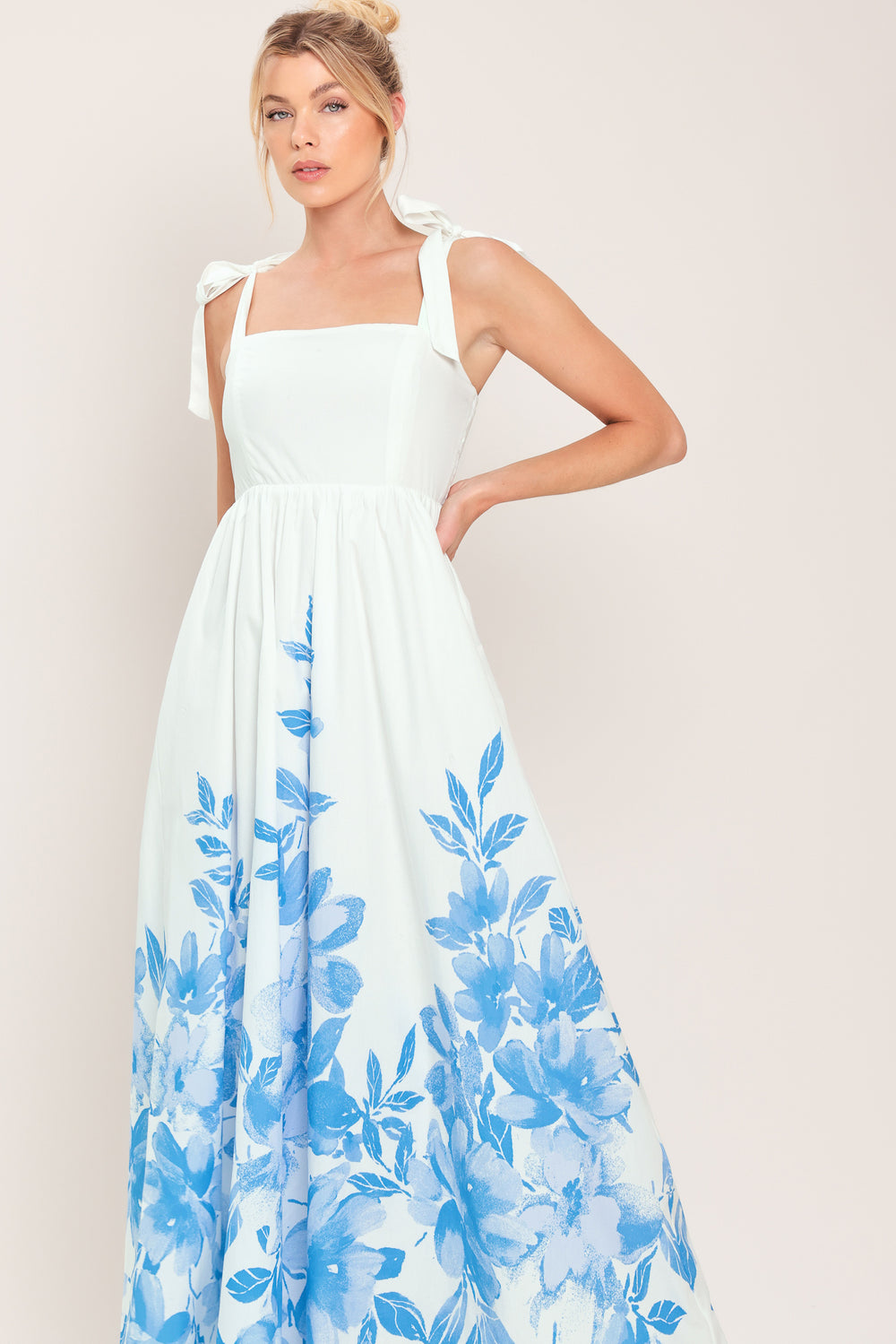 Woman wearing a white maxi dress with blue floral pattern featuring straight neckline, shoulder ties, full skirt and smocked back bodice | Flying Tomato