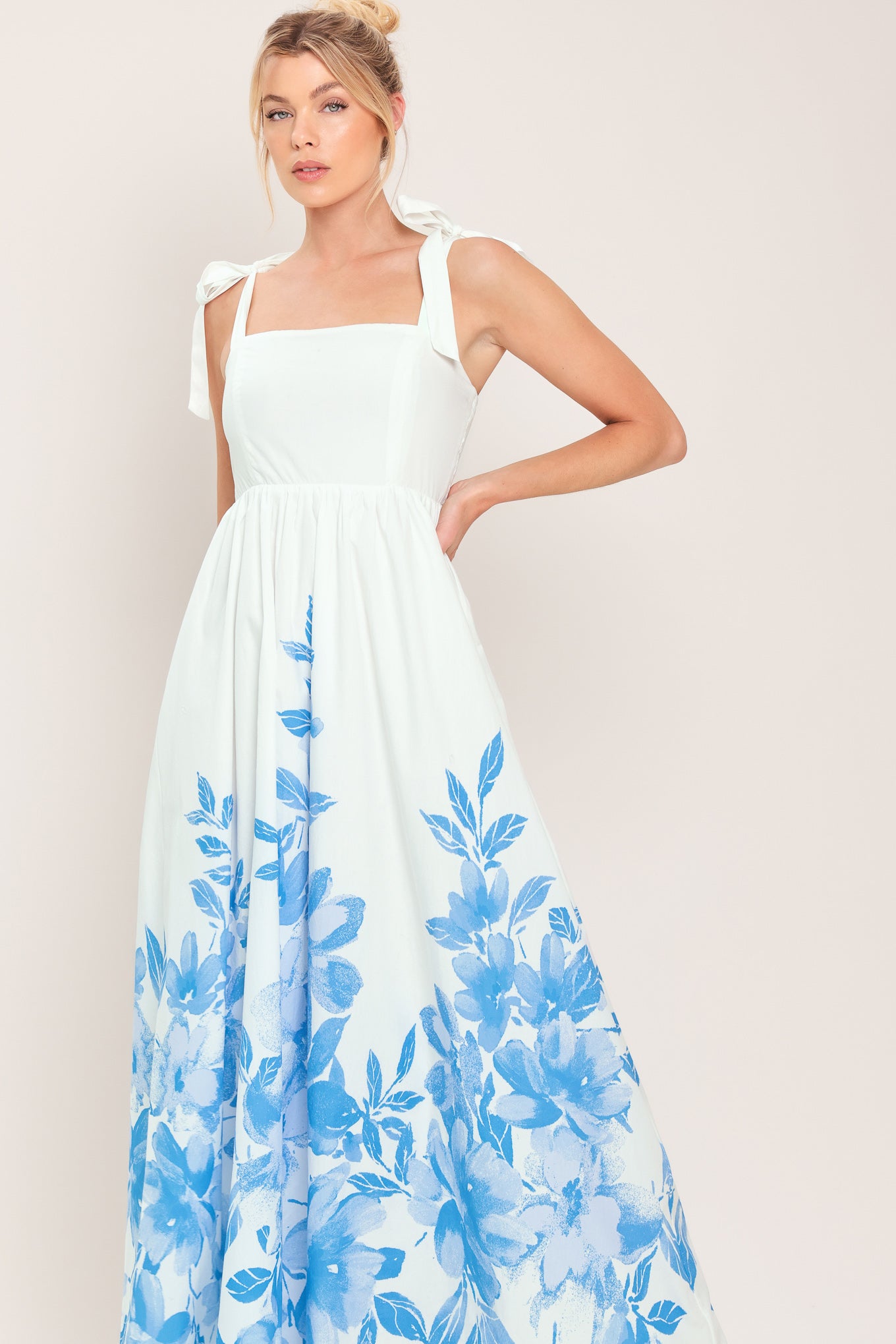 Woman wearing a white maxi dress with blue floral pattern featuring straight neckline, shoulder ties, full skirt and smocked back bodice | Flying Tomato