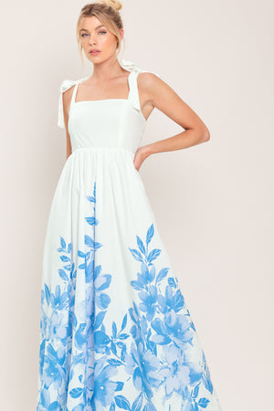 Woman wearing a white maxi dress with blue floral pattern featuring straight neckline, shoulder ties, full skirt and smocked back bodice | Flying Tomato