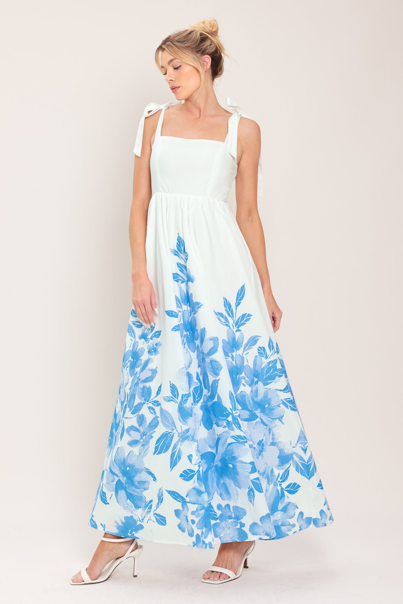 Woman wearing a white maxi dress with blue floral pattern featuring straight neckline, shoulder ties, full skirt and smocked back bodice | Flying Tomato